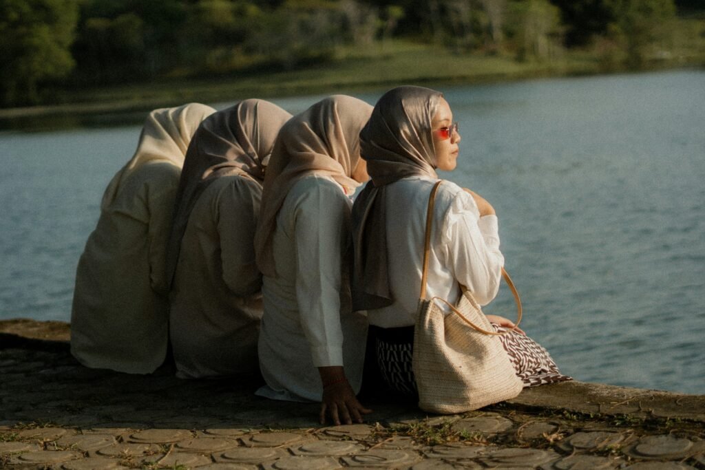 Four Muslim women in hijabs sit by a lake, one facing forward with sunglasses, symbolizing individuality within community.
