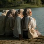 Four Muslim women in hijabs sit by a lake, one facing forward with sunglasses, symbolizing individuality within community.