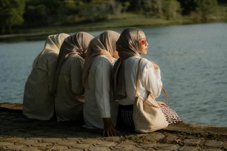 Four Muslim women in hijabs sit by a lake, one facing forward with sunglasses, symbolizing individuality within community.