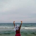 Wanda in a grey hijab and red dress stands on Jumunjin Beach, Gangneung-si (circa 2019), facing the sea with arms lifted high—half-playfully “summoning Cthulhu,” half-celebrating her own hard-won freedom and self-worth beneath an overcast sky and rolling waves.