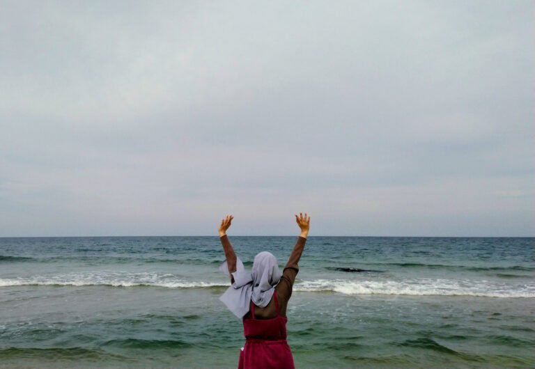 Wanda in a grey hijab and red dress stands on Jumunjin Beach, Gangneung-si (circa 2019), facing the sea with arms lifted high—half-playfully “summoning Cthulhu,” half-celebrating her own hard-won freedom and self-worth beneath an overcast sky and rolling waves.