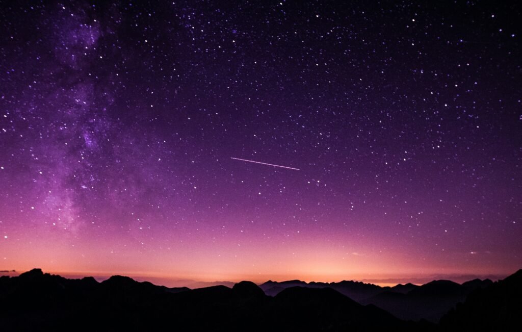Vast purple night sky scattered with stars above shadowed mountains, crossed by a faint shooting star.