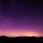 Vast purple night sky scattered with stars above shadowed mountains, crossed by a faint shooting star.