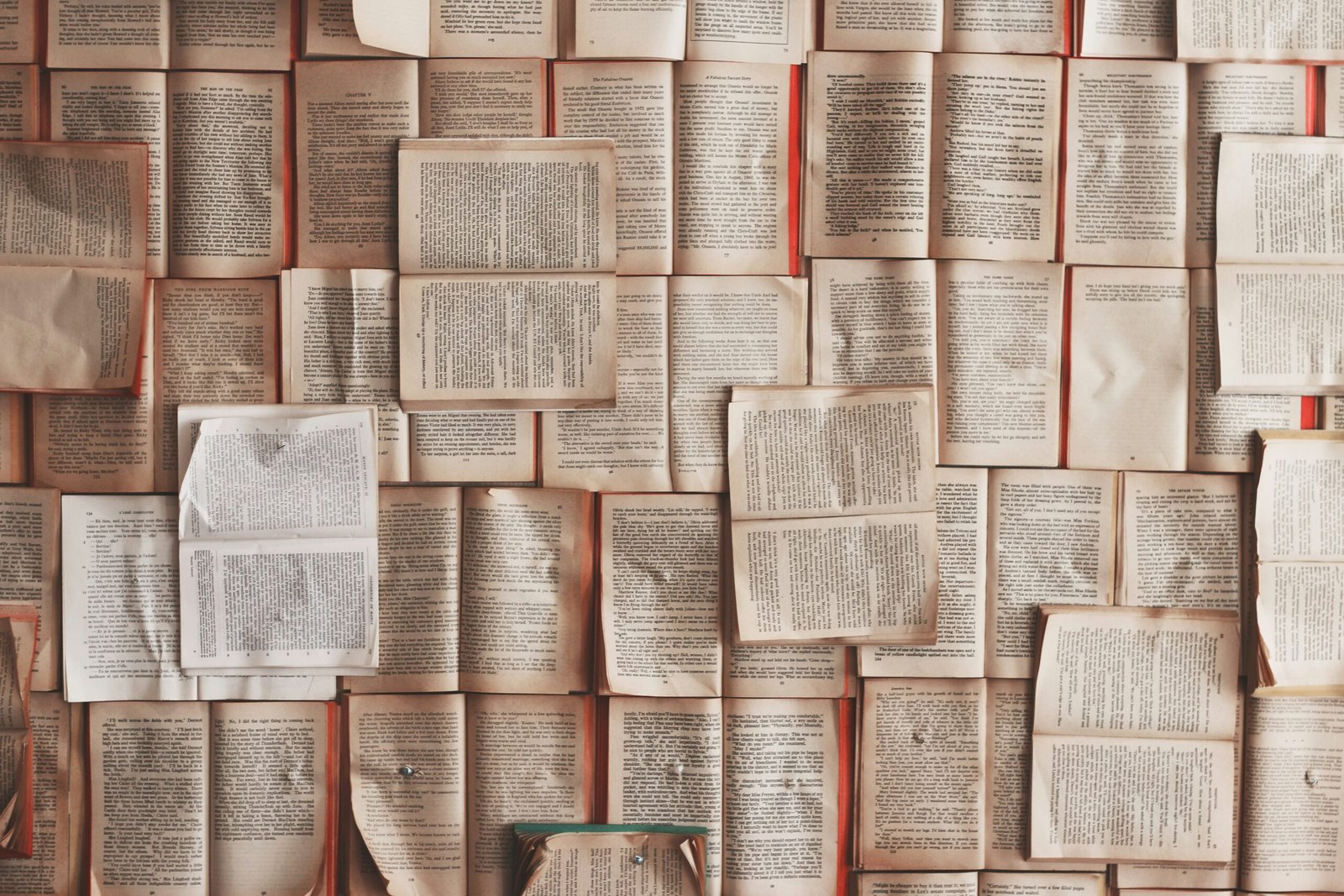 A top-down view of dozens of open books and loose pages arranged tightly together, forming a layered collage of printed text in warm, muted tones.