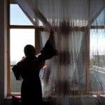 A woman photographed from behind in silhouette stands before a large sunlit window, one arm raised to gather the folds of a sheer embroidered curtain. Her figure is cast entirely in shadow against the brightness outside. Photograph by Wakil Kohsar/AFP/Getty Images.