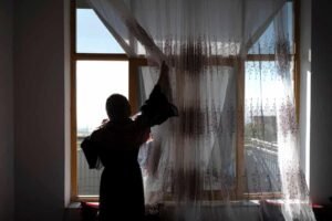 A woman photographed from behind in silhouette stands before a large sunlit window, one arm raised to gather the folds of a sheer embroidered curtain. Her figure is cast entirely in shadow against the brightness outside. Photograph by Wakil Kohsar/AFP/Getty Images.