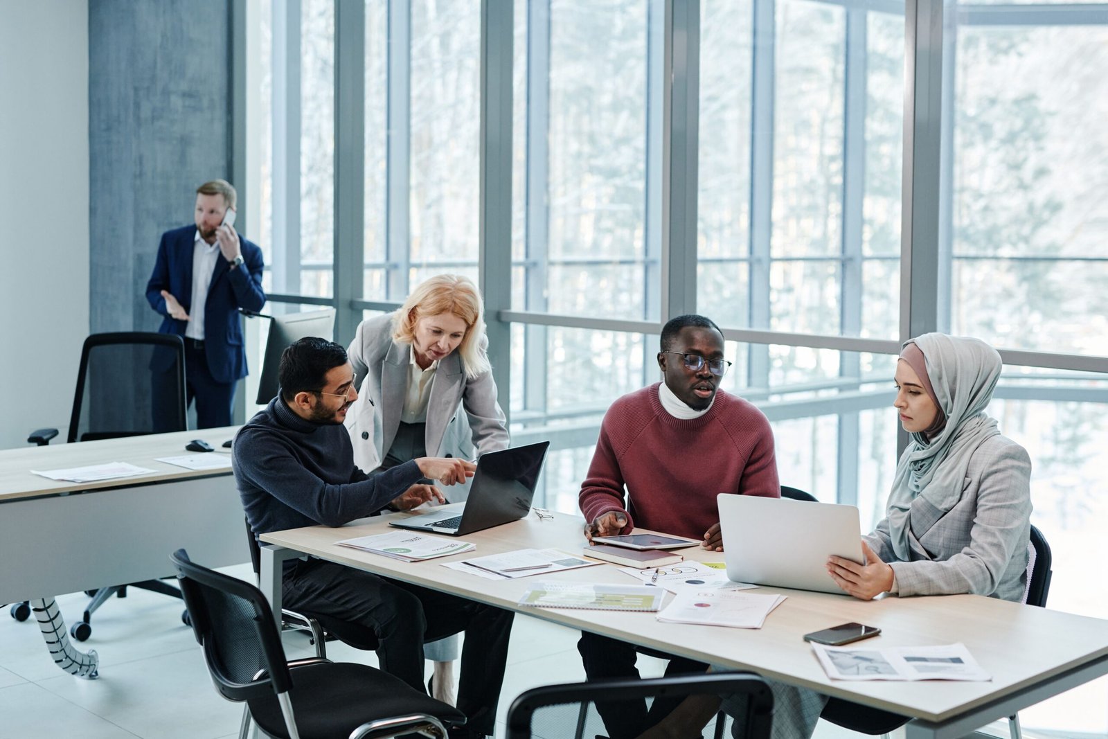 Five colleagues in a modern office: a woman in a light grey hijab works on a laptop beside a man in a burgundy sweater, while another man and a blonde woman discuss a second laptop and a suited man speaks on the phone in the background.