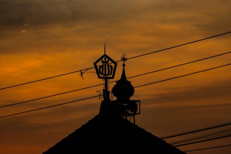 Silhouette of a mosque roof, dome, loudspeaker, and Allah ornament against a deep orange sunset sky, crossed by overhead power lines.