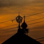 Silhouette of a mosque roof, dome, loudspeaker, and Allah ornament against a deep orange sunset sky, crossed by overhead power lines.