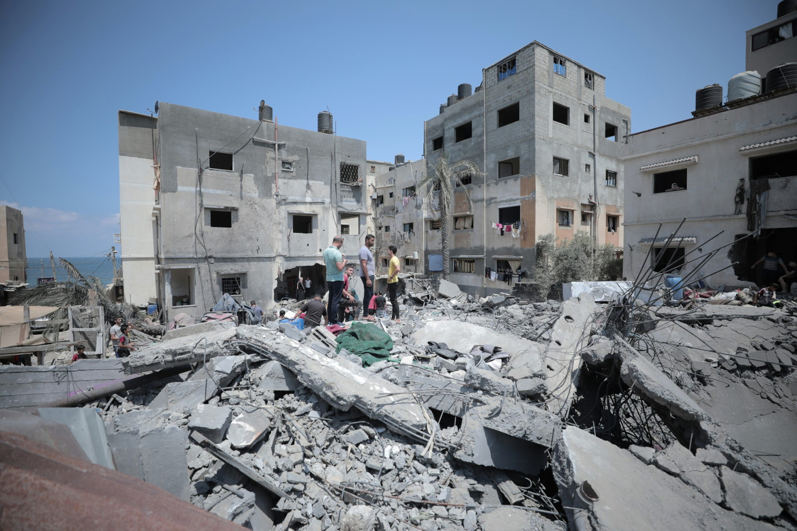 People stand amid the rubble of destroyed buildings in Gaza, surrounded by collapsed concrete slabs, exposed steel reinforcement, and heavily damaged residential buildings under a clear blue sky.