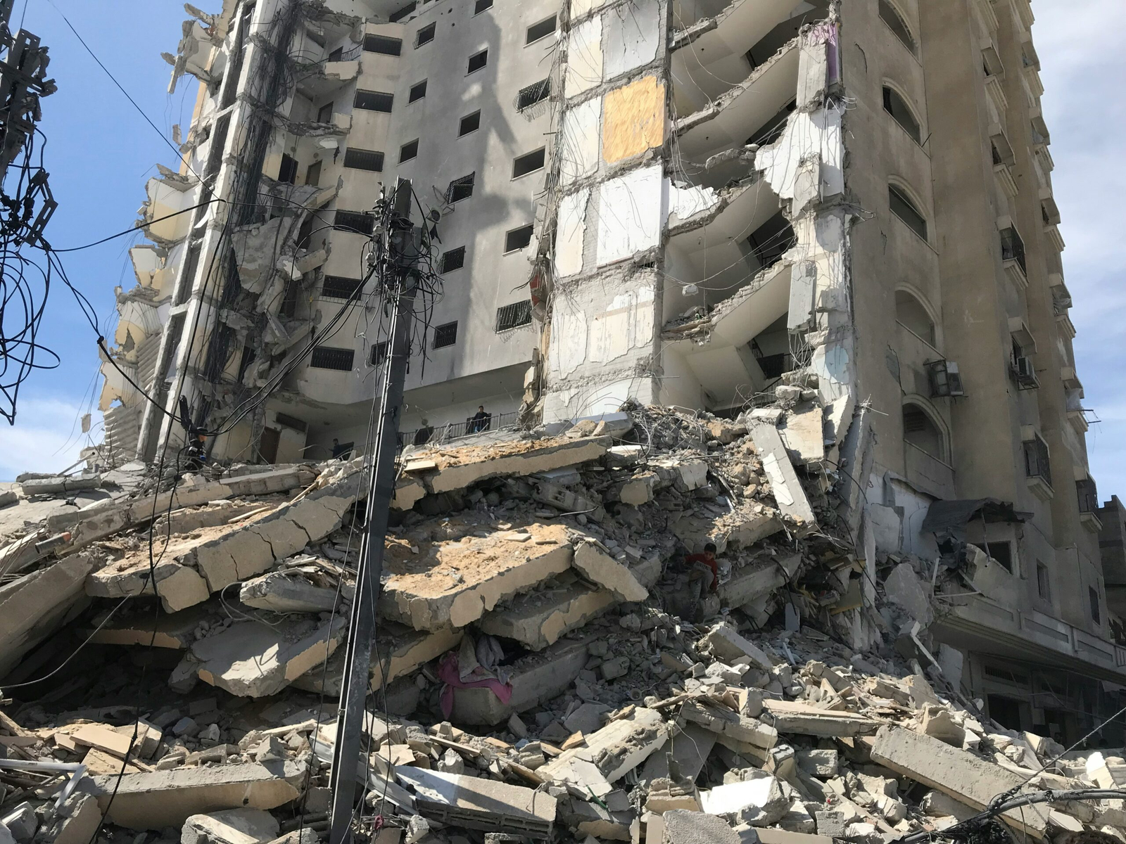 A heavily damaged multistorey building in Gaza with collapsed concrete floors, exposed steel reinforcement, and piles of rubble. Electrical poles and tangled cables stand in front of the ruins.