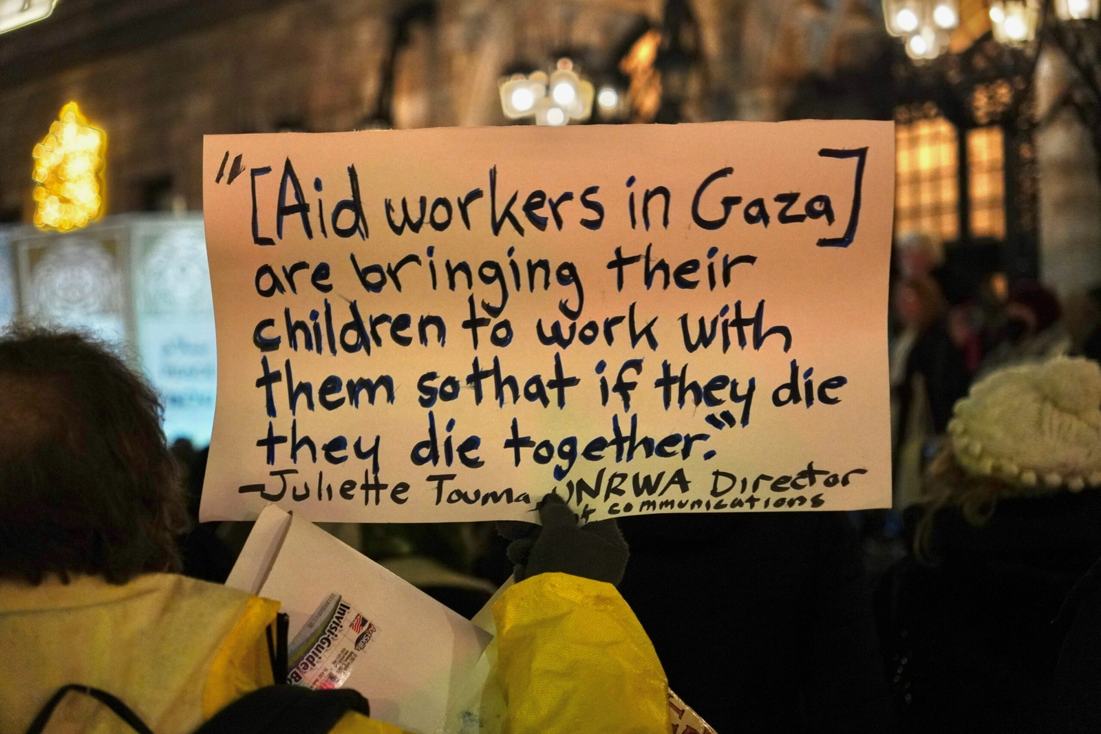 A protester holds a handwritten cardboard sign at a nighttime demonstration reading, "[Aid workers in Gaza] are bringing their children to work with them so that if they die they die together." The quote is attributed to Juliette Touma, UNRWA Director of Communications.